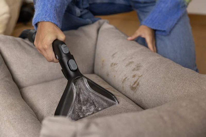 Person reinigt graues Sofa mit Kärcher Polsterreiniger. Flecken auf dem Stoff sichtbar. Jeans und blauer Pullover im Hintergrund.