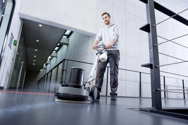 Person bedient Kärcher Bodenreinigungsmaschine in modernem, lichtdurchflutetem Gebäude mit Betonwänden und Geländer.