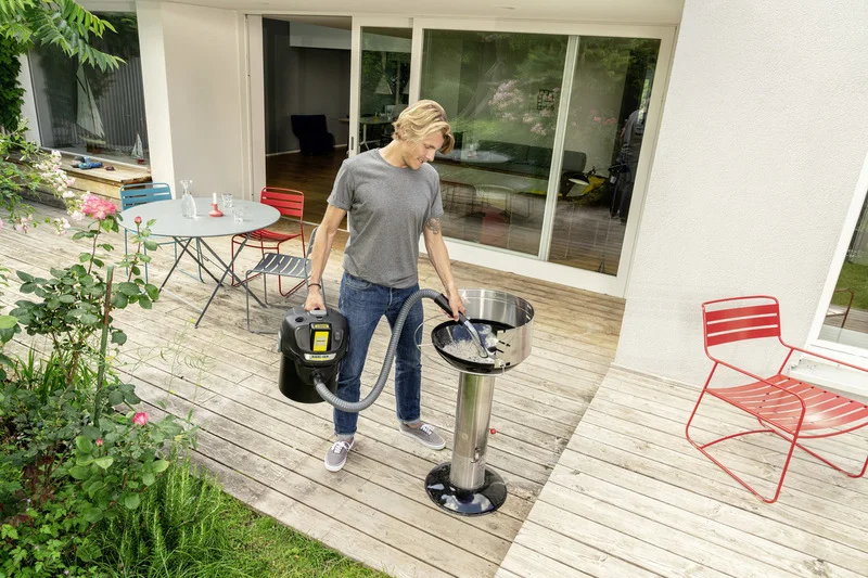 Person auf Terrasse nutzt Kärcher Aschesauger, um Grill zu reinigen. Tisch mit Glas und Stühlen im Hintergrund, Pflanzen seitlich.