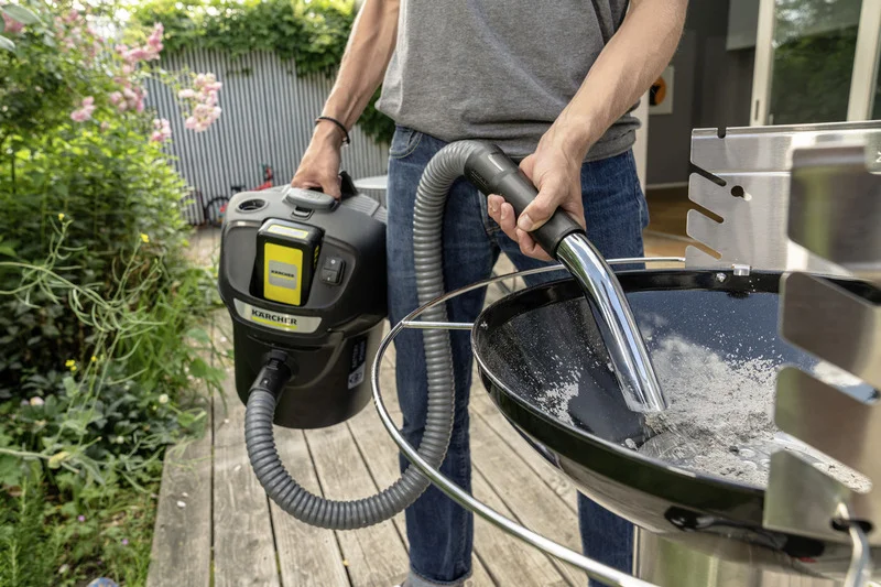 Person reinigt Grill mit Kärcher Aschesauger im Garten. Holzterrasse, Pflanzen im Hintergrund.
