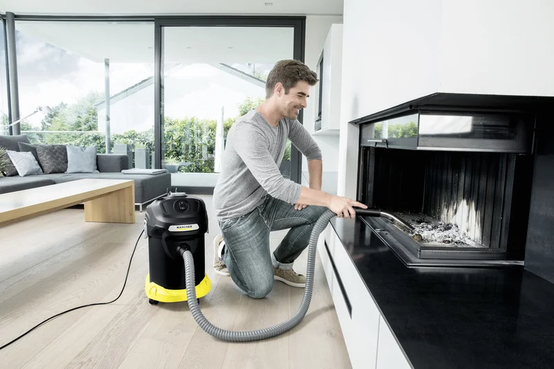 Person reinigt Kamin mit Kärcher Aschesauger in modernem Wohnzimmer mit großen Fenstern und hellen Möbeln.