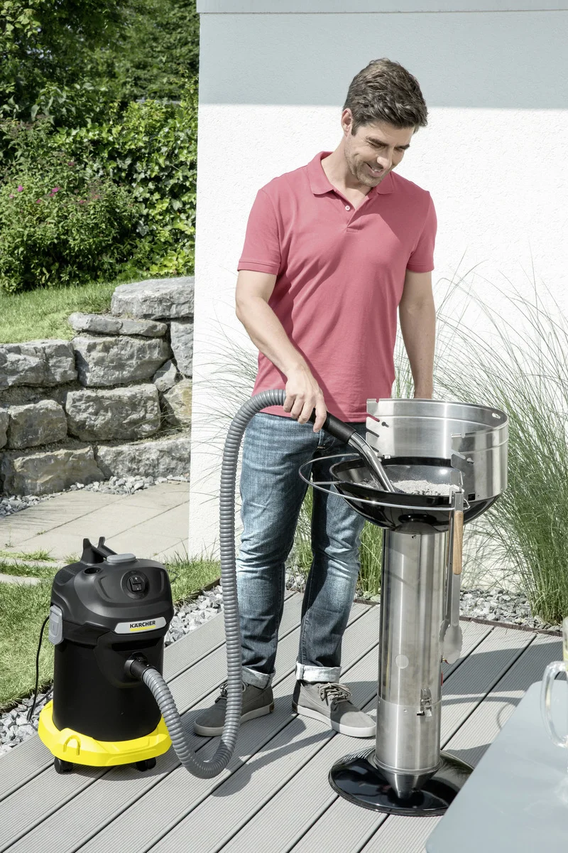 Mann in rotem Shirt reinigt Grill mit Kärcher Aschesauger im Garten. Hintergrund mit Steinmauer und Pflanzen.