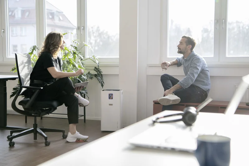 Zwei Personen sitzen in einem hellen Büro, eine auf einem Stuhl, die andere auf einer Fensterbank. Ein Luftreiniger steht zwischen ihnen.