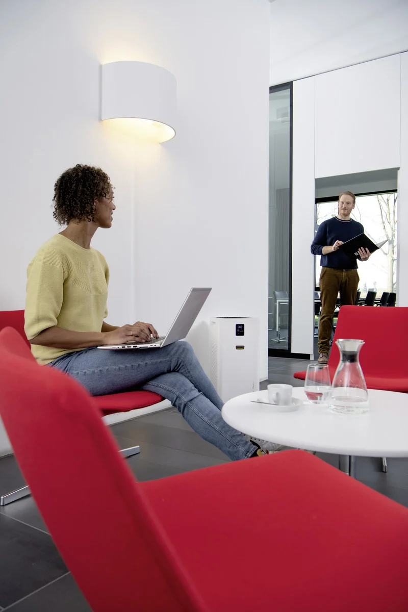 Frau sitzt mit Laptop auf rotem Stuhl in modernem Büro, neben einem Tisch mit Karaffe und Tassen. Im Hintergrund steht ein Mann mit Notizbuch.