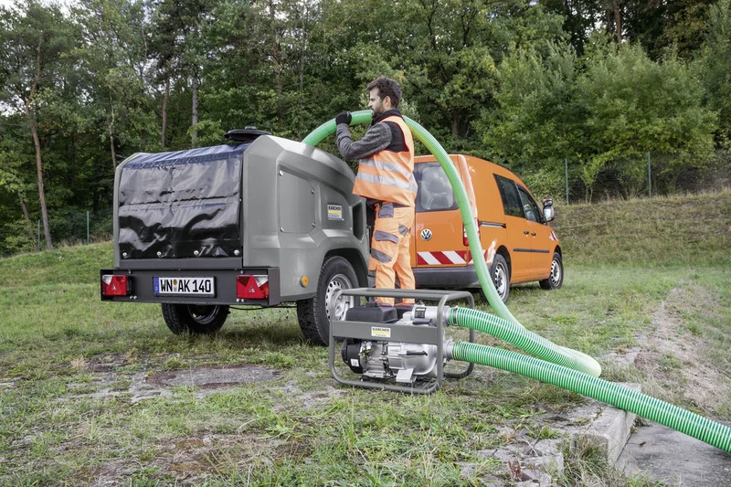 Person in orange Arbeitskleidung bedient Kärcher Wasserpumpe im Freien, neben einem grauen Anhänger und einem orangefarbenen VW Caddy.