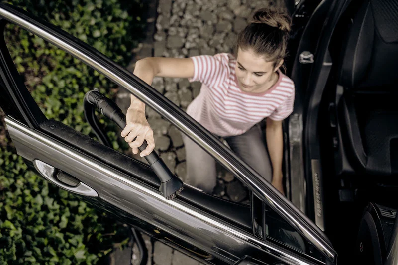 Person reinigt Autotür mit einem Staubsauger. Auto steht auf gepflastertem Boden, umgeben von grünen Pflanzen.