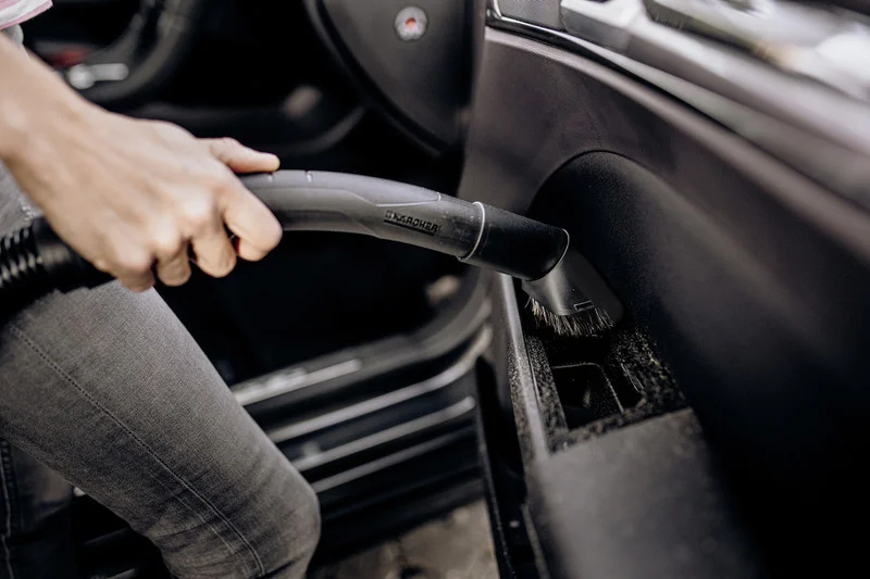 Hand hält Kärcher Staubsaugerdüse, reinigt Autoinnenraum. Graue Jeans sichtbar, Fokus auf Staubsauger und Armaturenbrett.