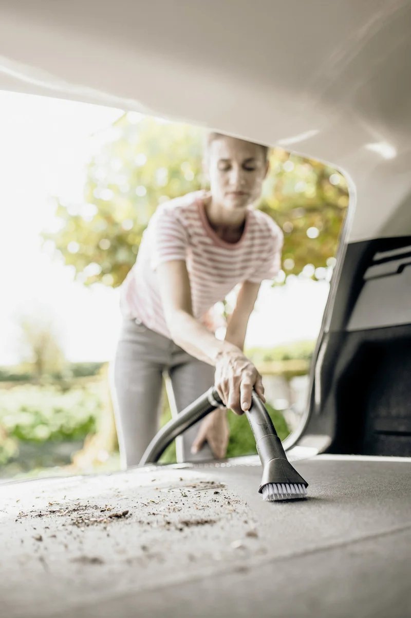 Person reinigt den Kofferraum eines Autos mit einem Staubsauger. Draußen, sonniges Wetter, Bäume im Hintergrund.