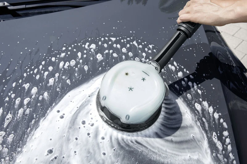 Hand hält Kärcher Autowaschbürste, reinigt schwarze Motorhaube mit Schaum und Wasser im Freien.