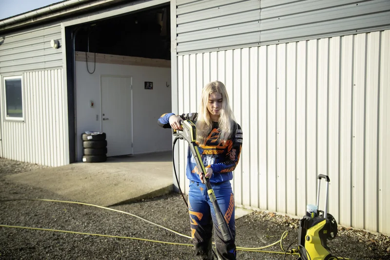 Person in Motorradkleidung reinigt im Freien mit einem Kärcher Hochdruckreiniger vor einer Garage. Reifenstapel im Hintergrund.