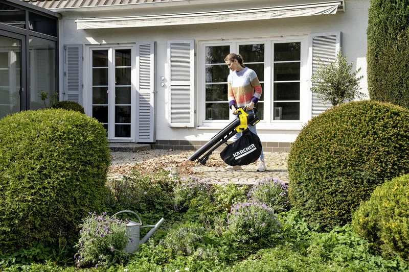 Person im Garten verwendet Kärcher Laubsauger, um Blätter vor einem Haus mit weißen Fensterläden zu entfernen.