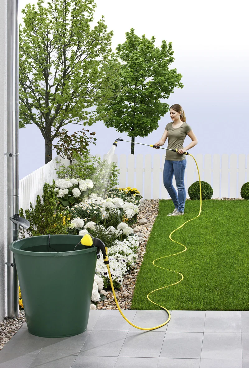 Frau bewässert Garten mit gelbem Schlauch. Grüner Eimer und Kärcher Gartenpumpe im Vordergrund. Blühende Pflanzen und Bäume im Hintergrund.
