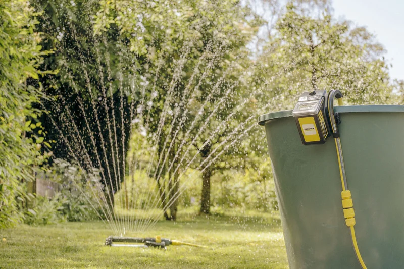 Kärcher Bewässerungssteuerung an grünem Fass, im Garten mit Sprinkler und Wasserstrahlen.