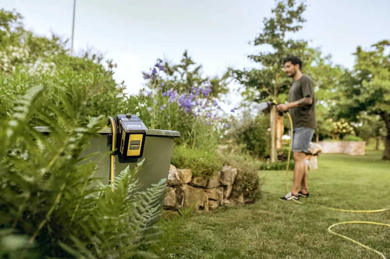 Mann bewässert Garten mit Schlauch, im Vordergrund ein Kärcher Bewässerungssteuergerät an einer Mülltonne.