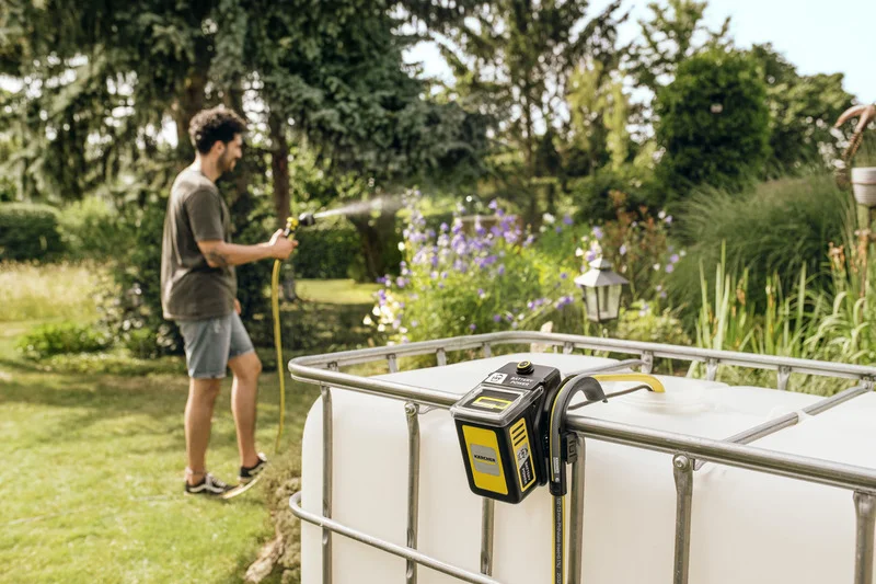 Mann bewässert Garten mit Schlauch, im Vordergrund ein Kärcher Bewässerungssteuergerät an einem Wassertank.