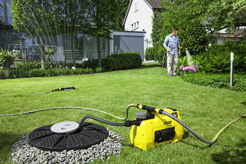 Mann bewässert Garten mit Kärcher Gartenpumpe, umgeben von grüner Rasenfläche und Blumenbeeten, im Hintergrund ein Haus.
