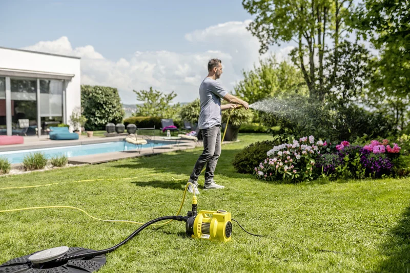 Mann bewässert Blumen im Garten mit einem Schlauch. Im Vordergrund steht eine gelbe Kärcher Gartenpumpe. Im Hintergrund ein Pool und Sitzmöbel.