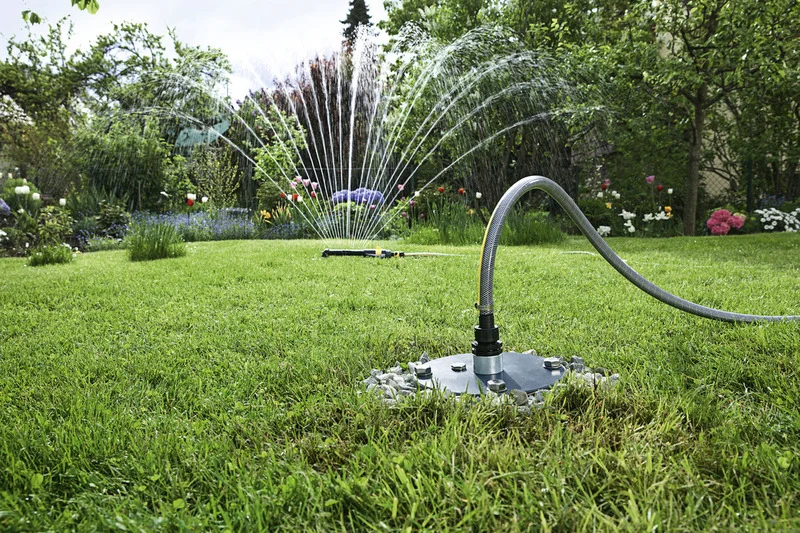 Gartenbewässerung mit Sprinkler, Wasserstrahlen formen Halbkreis, grüner Rasen, blühende Pflanzen im Hintergrund.