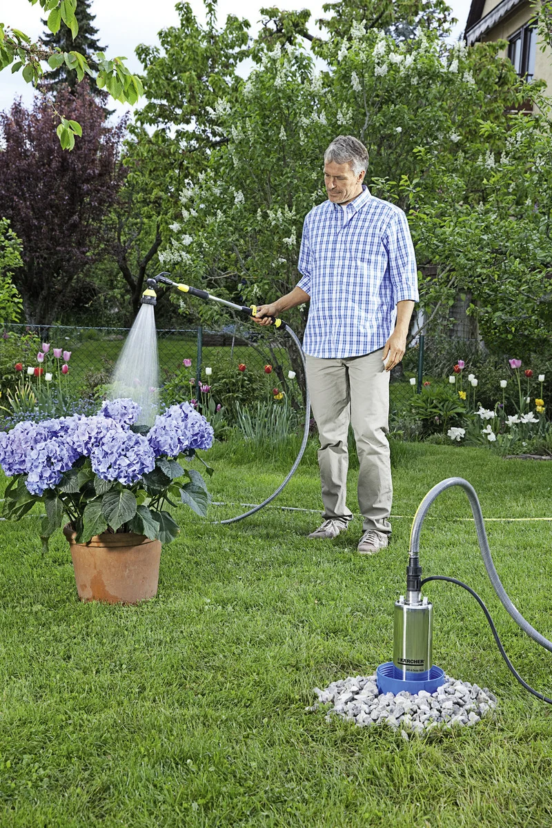 Mann bewässert Hortensien im Garten mit einem Schlauch, verbunden mit einer Kärcher Gartenpumpe, umgeben von blühenden Pflanzen.