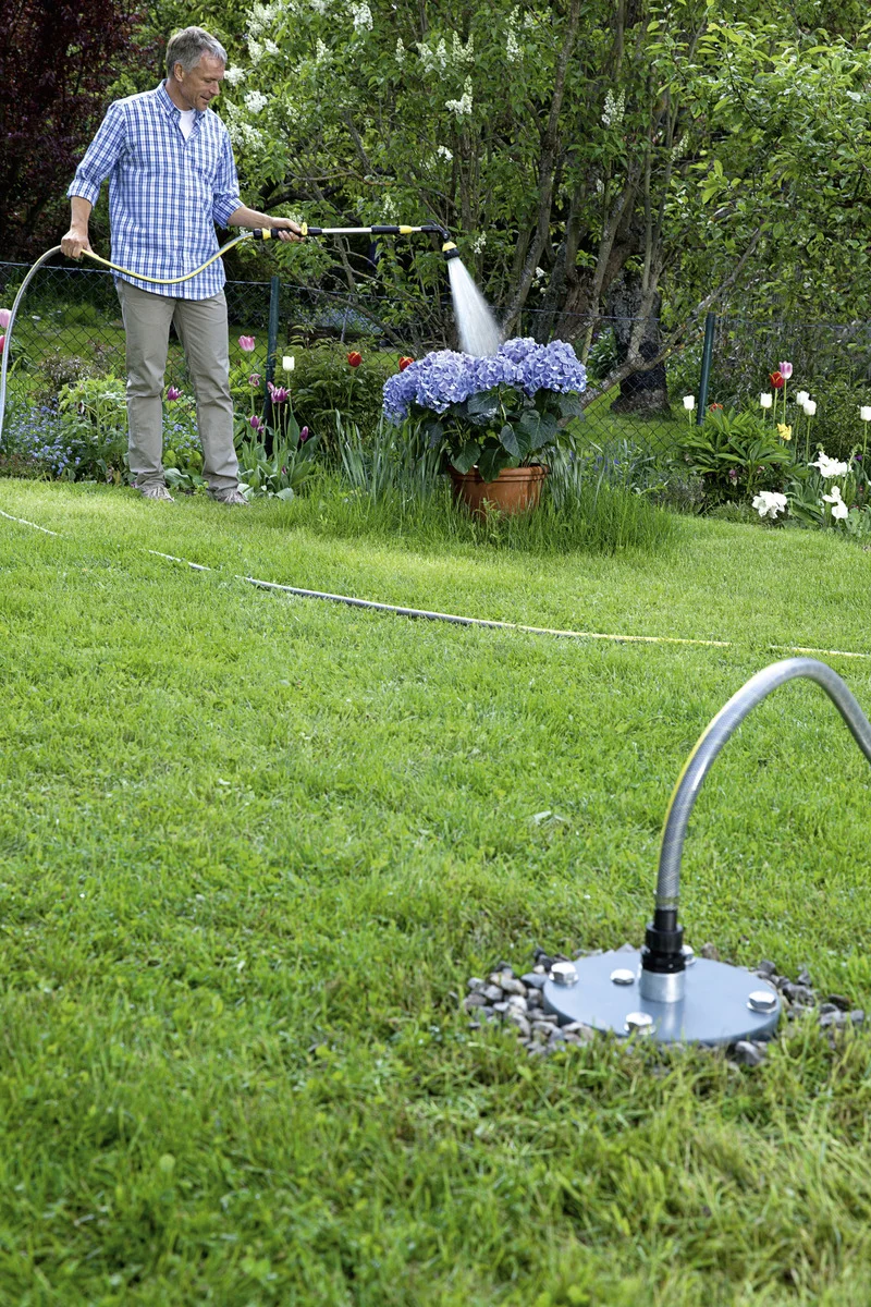 Mann bewässert Hortensien im Garten mit einem Schlauch. Im Vordergrund ein Wasseranschluss auf grünem Rasen.