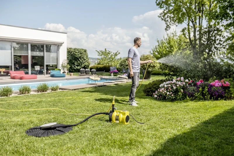 Mann bewässert Garten mit Schlauch neben einem Pool. Im Vordergrund ein gelber Kärcher Gartenpumpe. Terrasse mit Sitzmöbeln im Hintergrund.