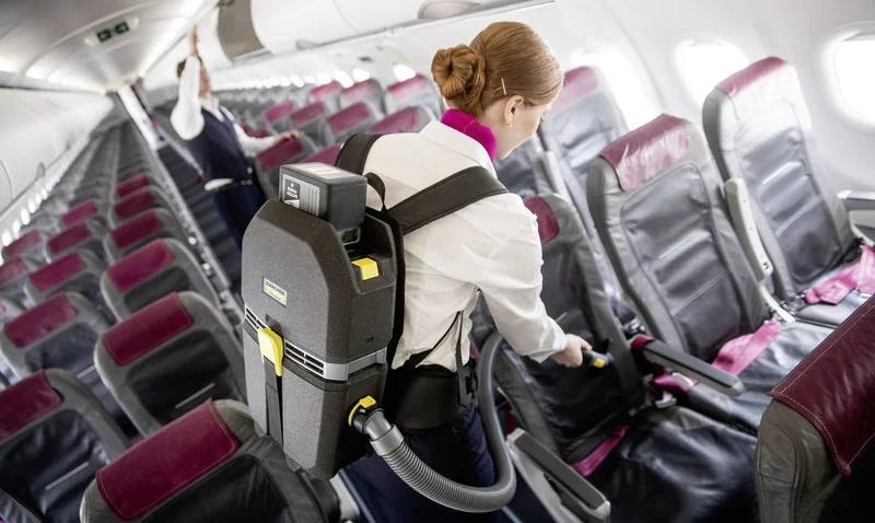 Person reinigt Flugzeugsitze mit einem Kärcher Rucksack-Staubsauger. Sitzreihen mit roten Kopfstützen in einem Flugzeug.