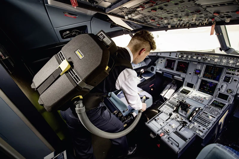 Person im Cockpit eines Flugzeugs mit einem Kärcher Rucksack-Staubsauger, umgeben von Instrumententafeln und Bedienelementen.