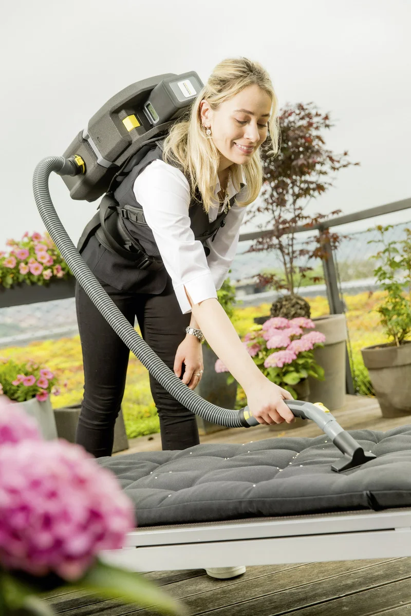 Frau reinigt Outdoor-Matratze mit einem Kärcher Rucksack-Staubsauger, umgeben von blühenden Pflanzen auf einer Terrasse.