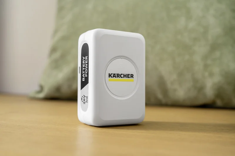 Weißer Kärcher Akku auf einem Holztisch, im Hintergrund ein grünes Kissen.