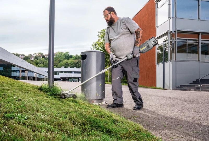 Person trimmt Gras mit einem Kärcher Rasentrimmer neben einem Mülleimer auf einem Gehweg vor einem modernen Gebäude.