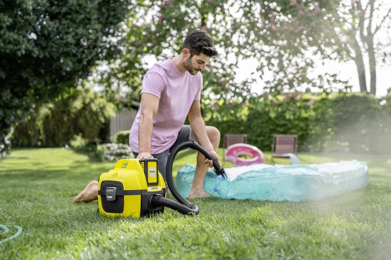 Mann in rosa T-Shirt bläst mit einem Kärcher Staubsauger ein aufblasbares Pool im Garten auf. Hintergrund mit Bäumen und Gartenmöbeln.