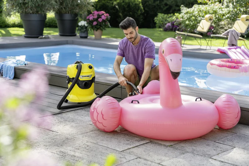 Mann füllt rosa Flamingo-Schwimmring mit Kärcher Nass-/Trockensauger auf, neben einem Pool im Garten. Im Hintergrund blühende Pflanzen.