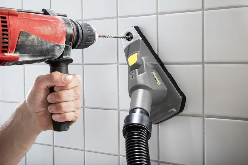 Hand hält Bohrmaschine, bohrt in Fliesenwand. Kärcher Staubabsaugung befestigt, graues Gerät mit gelbem Detail.