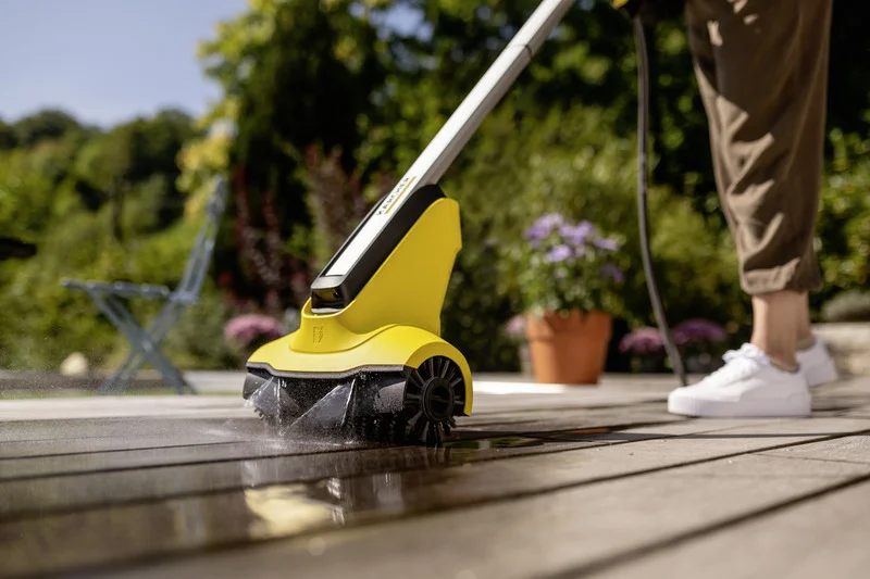Kärcher Terrassenreiniger reinigt Holzterrasse im Garten. Person in weißen Schuhen bedient das Gerät. Blumen im Hintergrund.