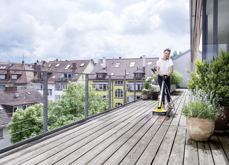 Person reinigt Holzterrasse mit Kärcher Bodenreiniger, umgeben von Pflanzen. Im Hintergrund sind mehrstöckige Wohnhäuser zu sehen.
