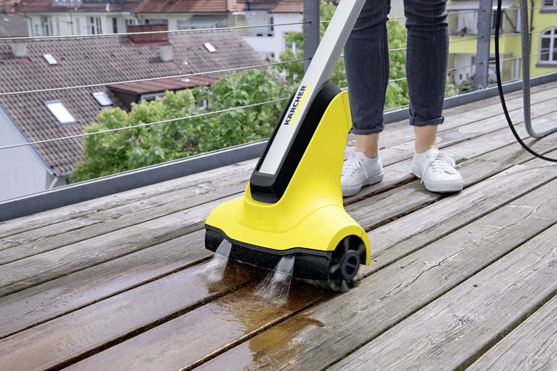 Person reinigt Holzboden mit Kärcher Bodenreiniger auf Balkon, umgeben von grünen Bäumen und Häusern im Hintergrund.