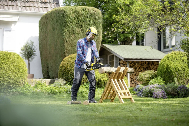 Person mit Schutzhelm und Handschuhen benutzt eine Kettensäge im Garten, um Holz auf einem Sägebock zu schneiden. Im Hintergrund sind Büsche und ein Holzlager.