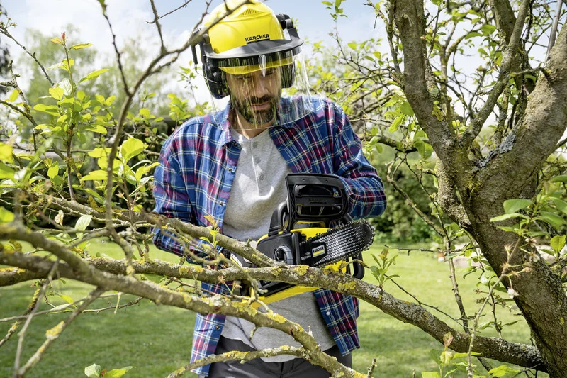Person mit Schutzhelm und Kärcher Kettensäge schneidet Äste im Garten. Umgeben von grünen Bäumen und Gras.