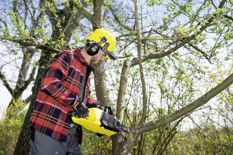 Person in kariertem Hemd und gelbem Schutzhelm schneidet mit einer Kärcher Akku-Kettensäge einen Ast im Freien.