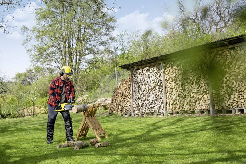 Mann mit Schutzhelm und Ohrenschützern schneidet Holz mit einer Kettensäge im Garten. Im Hintergrund gestapeltes Brennholz.