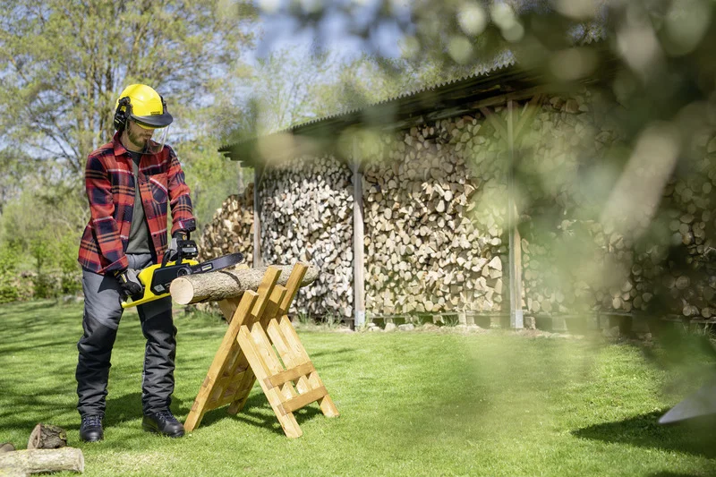 Person mit Schutzhelm und Kettensäge schneidet Holzscheit auf einem Holzgestell im Garten, im Hintergrund Holzstapel.