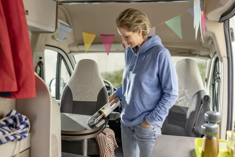 Person in blauem Hoodie reinigt Tisch im Wohnmobil mit einem Kärcher Handstaubsauger. Bunte Wimpel hängen von der Decke.