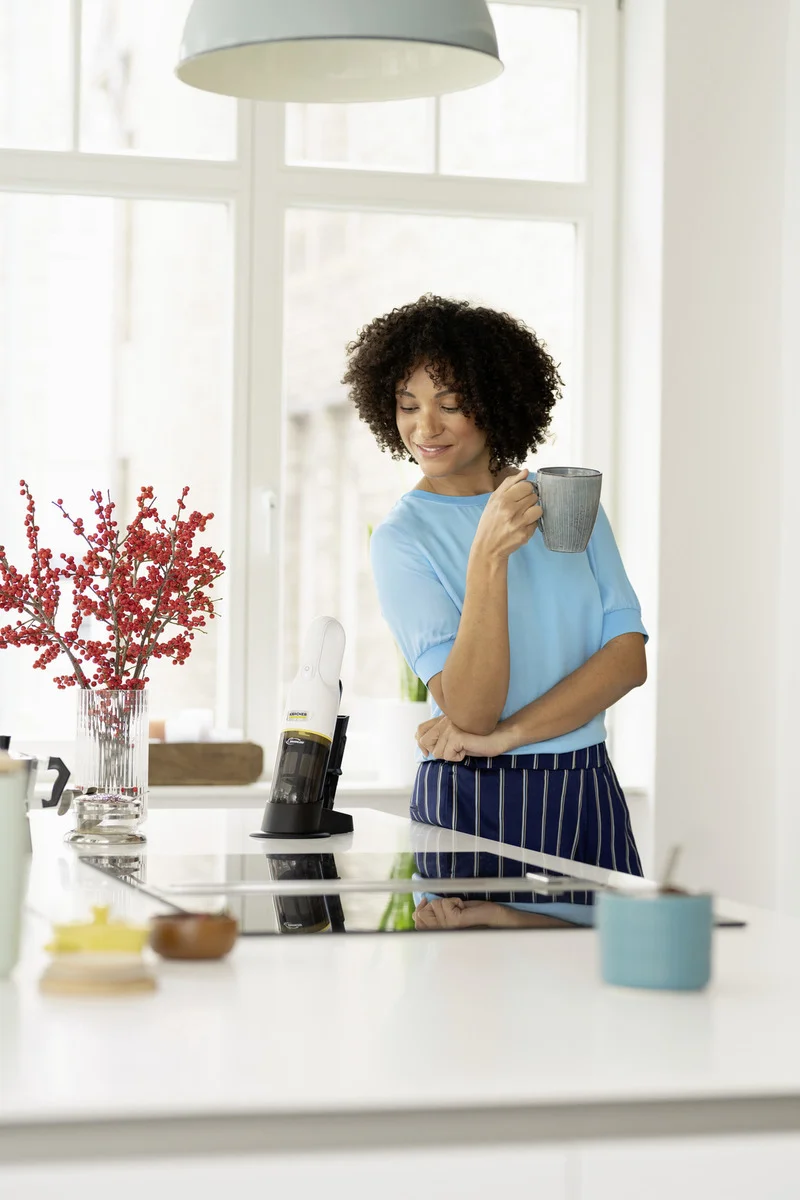Person in blauer Kleidung hält Tasse in moderner Küche. Ein Kärcher Fenstersauger steht auf der Arbeitsplatte neben roten Beeren in Vase.