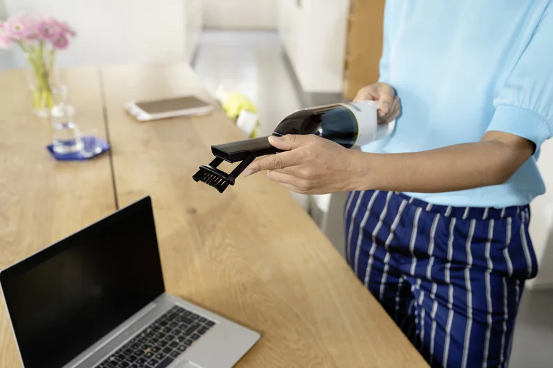 Person reinigt Laptop mit einem Kärcher Handstaubsauger auf einem Holztisch in einem hellen Raum.