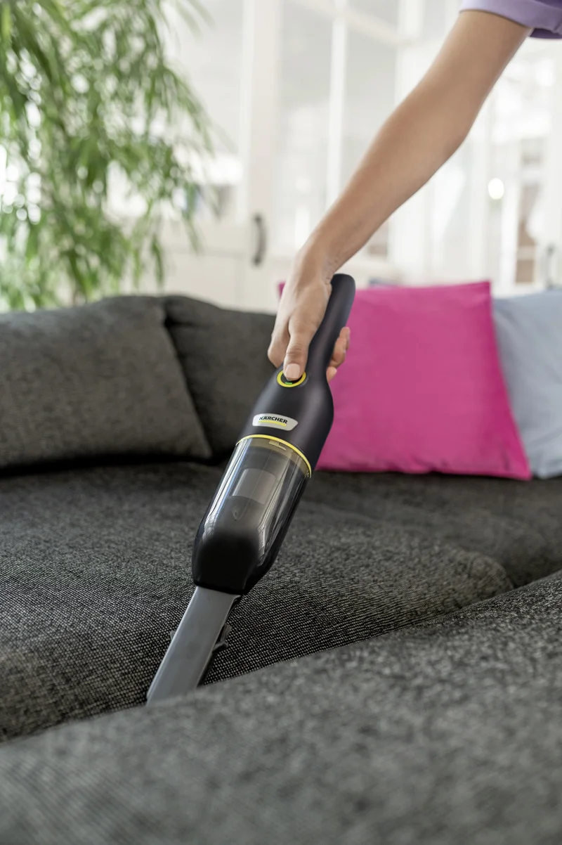 Hand hält Kärcher Handstaubsauger über grauem Sofa, im Hintergrund bunte Kissen und Pflanzen.