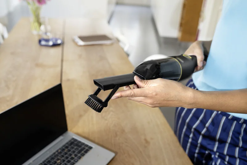 Person hält Kärcher Handstaubsauger mit Bürstenaufsatz über einem Laptop auf einem Holztisch in einem hellen Raum.