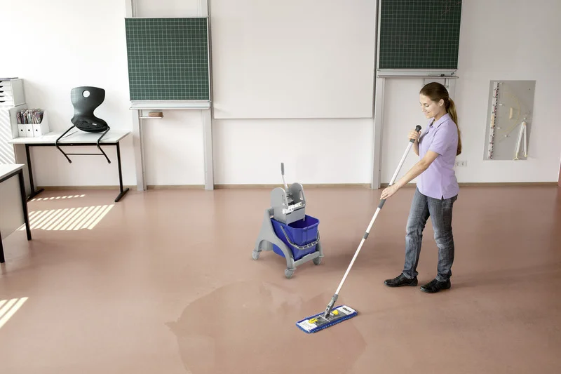 Person reinigt Boden in einem Klassenzimmer mit einem Wischmopp. Im Hintergrund sind Tafeln und ein Schreibtisch zu sehen.