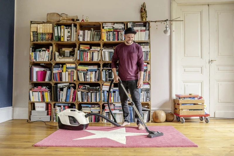 Person in rotem Pullover mit Staubsauger auf rosa Teppich vor Bücherregal in Wohnzimmer. Holzboden und weiße Tür im Hintergrund.