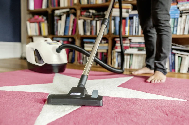 Person reinigt Teppich mit Kärcher Staubsauger in einem Raum mit Bücherregal im Hintergrund.