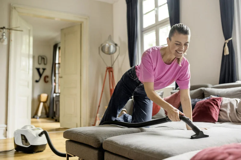 Person reinigt Sofa mit einem Kärcher Staubsauger in einem hellen Wohnzimmer mit Holzboden und großen Fenstern.